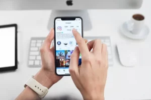 Person holding a smartphone displaying an Instagram profile on a white desk with a keyboard.