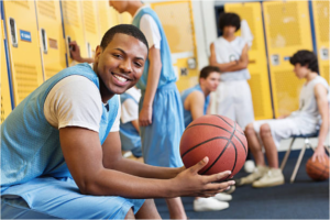 Smiling student-athlete holding a basketball in a locker room with teammates.
